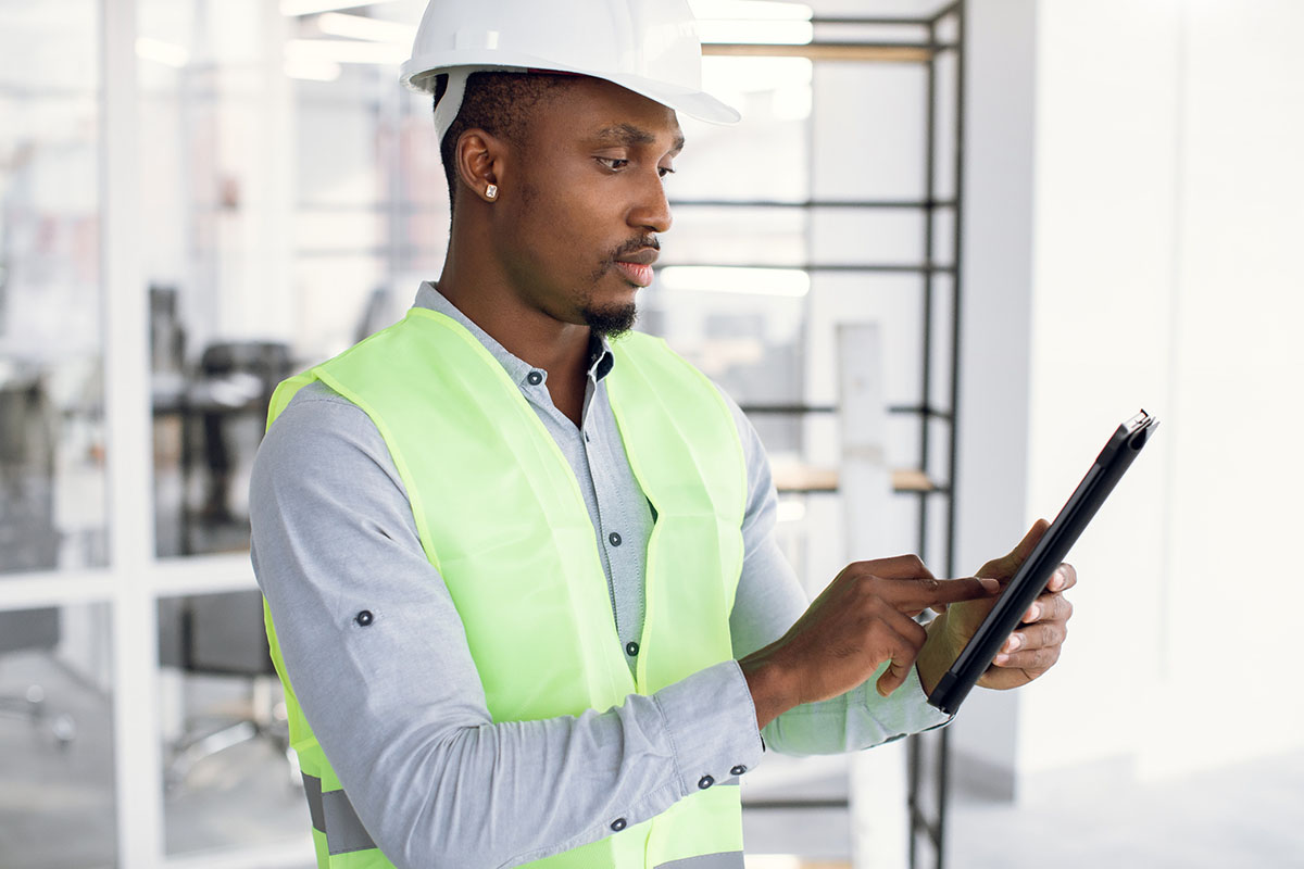 Focused inspector using digital tablet on construction object. Handsome man wearing uniform and white hardhat.